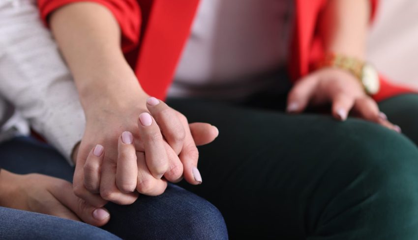 Two women tightly holding hands on couch at home close-up. Psychological support concept